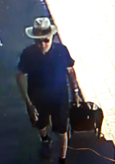 Man wearing cream wide brimmed hat, blue polo shirt, black shorts, pulling a black trolley