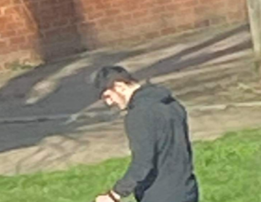 A man walking on a footpath. He is wearing a black hoodie. He has black hair. Behind him is a small patch of grass next to concrete. 