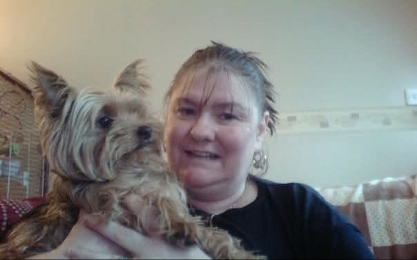 White woman, with tied back brown hair, smiling and holding a dog