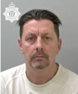 A man with short brown hair wearing a white shirt standing infront of a grey wall