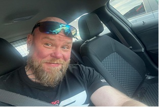 A bearded man with sunglasses on his head smiles in a car, wearing a black T-shirt. The interior is grey, conveying a casual and cheerful tone.