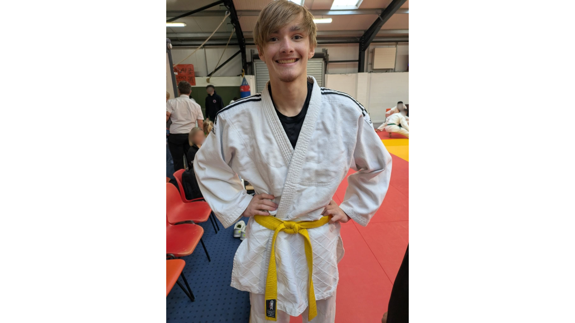 A smiling young man in a white judo gi and yellow belt standing in a martial arts dojo.
