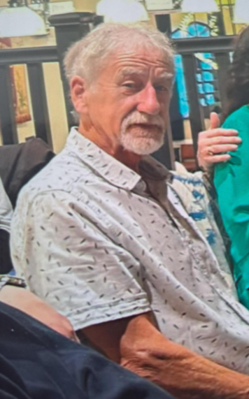 An older man with grey hair and a goatee, wearing a patterned light-coloured shirt, looking towards the camera.