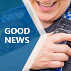 A close up of a female Police Community Support Officer who is holding their airwave radio on the right. They are wearing a blue undershirt. There is a blue arrow shape on the right with a white text on top that says ‘GOOD NEWS’